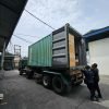 Kontainer kaca besar sedang dipindahkan dengan truk di area industri.