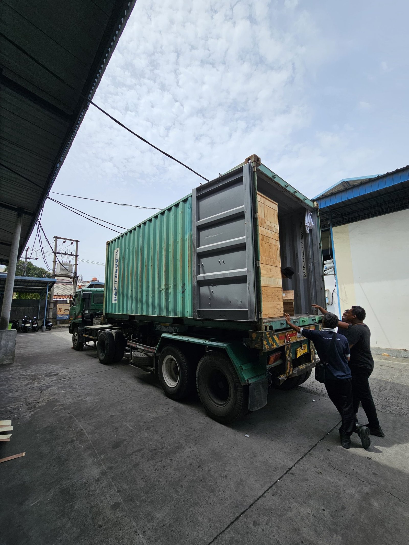 Kontainer kaca besar sedang dipindahkan dengan truk di area industri.